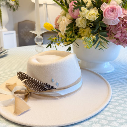 White hat with feather and ribbon on a table with floral arrangements in the background