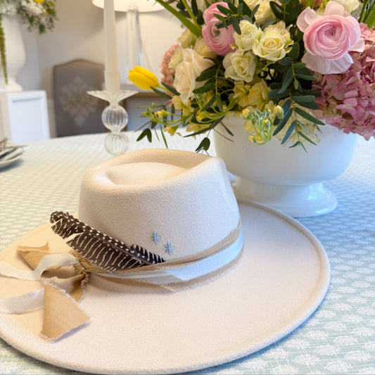 White hat with feather and ribbon on a table with floral arrangements in the background